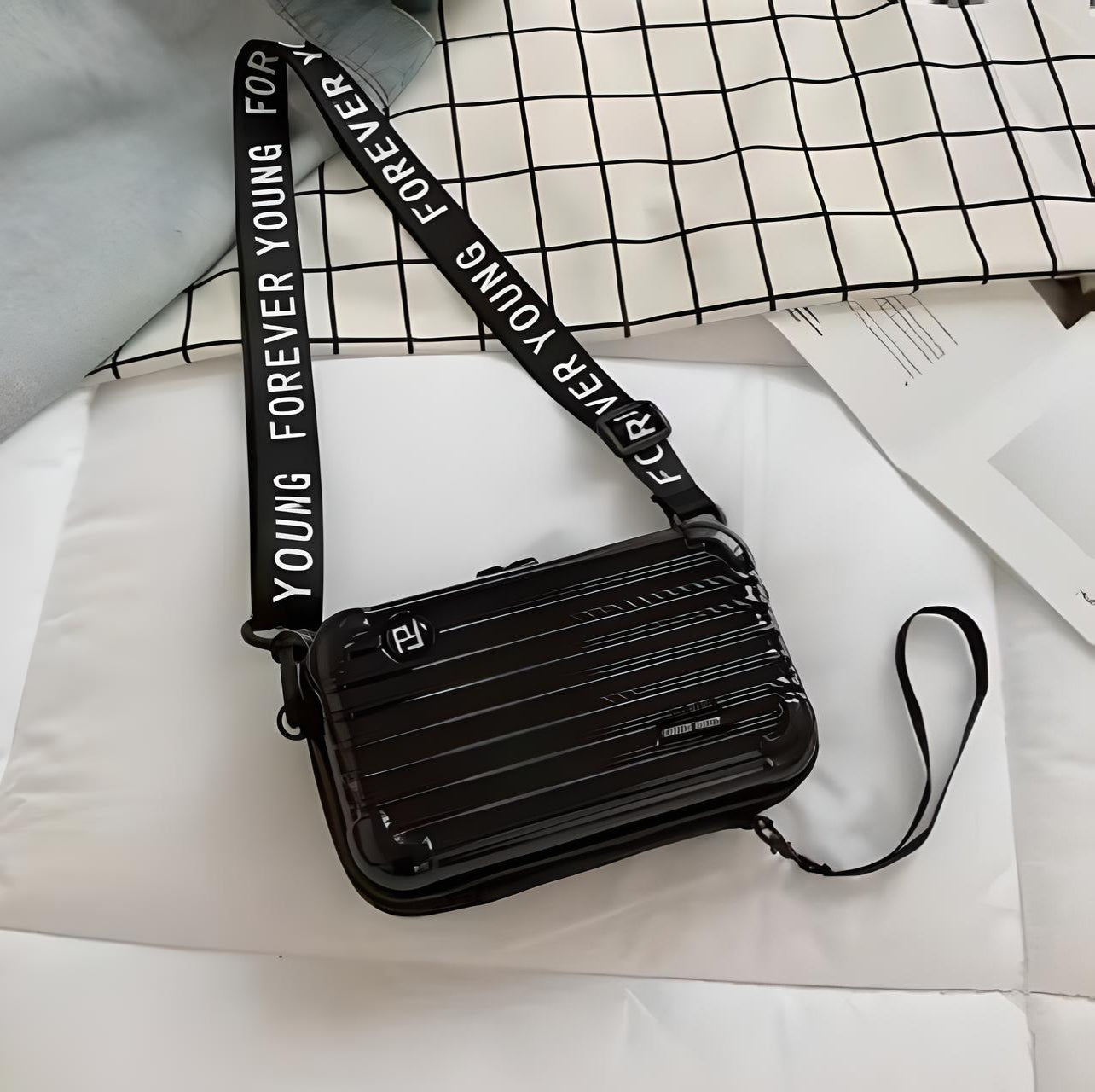 Black hard-shell crossbody bag with "Forever Young" adjustable strap on a checkered surface; stylish, durable, and modern accessory for everyday use.