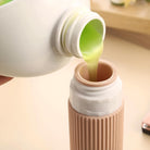 Pouring green liquid from a white bottle into a beige container, close-up. Skincare product transfer, beauty routine, eco-friendly packaging.