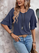 Woman wearing a stylish blue ruffle sleeve top with a boho necklace and jeans, leaning against a textured wall. Fashionable casual outfit.