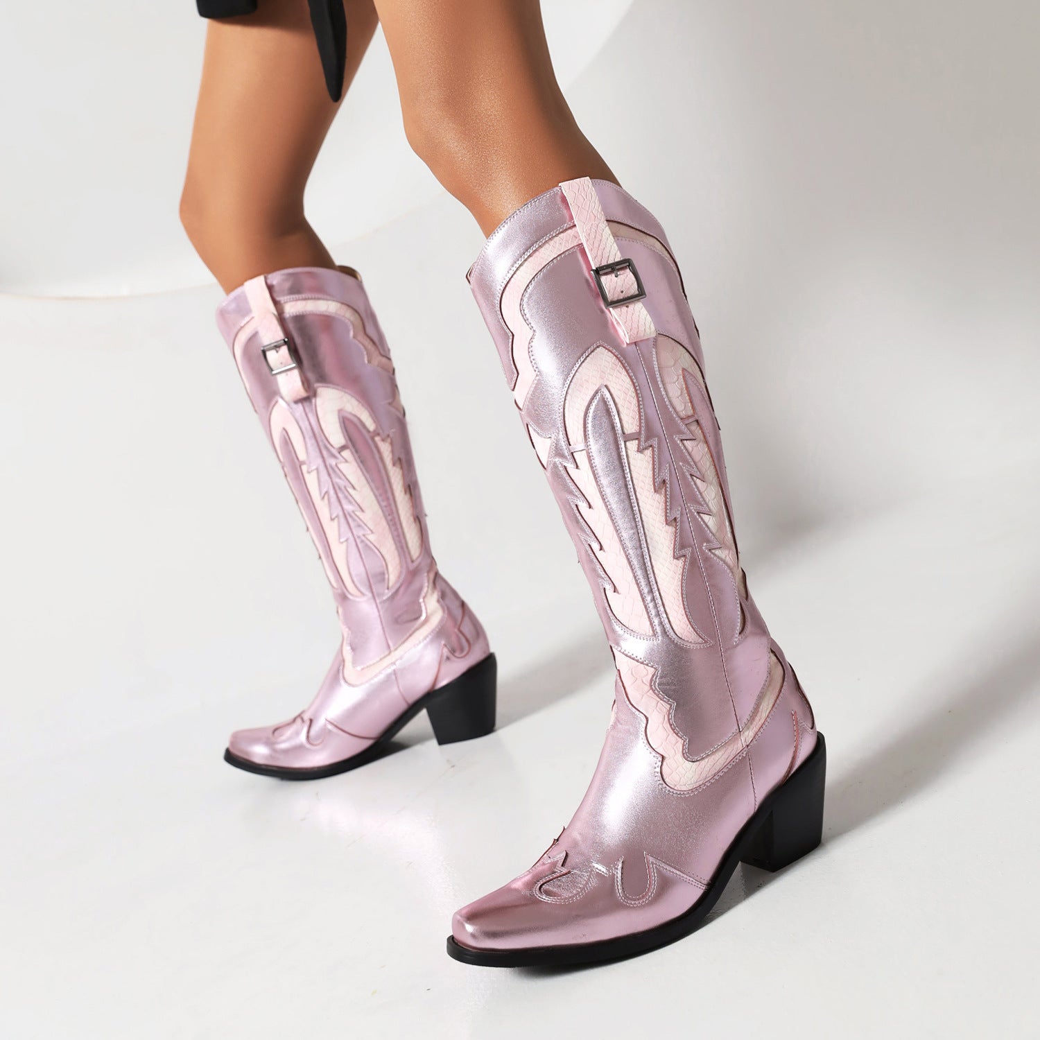 Pink metallic cowboy boots with intricate stitching and block heels, worn by a model. Fashionable western footwear, stylish women's boots.