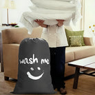 Laundry bag with "wash me" text and smiley face in a cozy living room setting, person holding folded linens. Perfect for home organization.