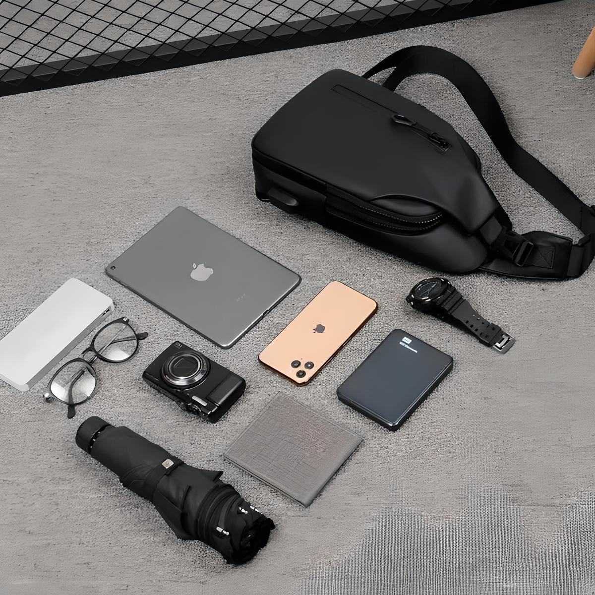 Flat lay of everyday carry essentials: black sling bag, iPad, iPhone, camera, glasses, umbrella, power bank, wallet, and smartwatch on a gray surface.