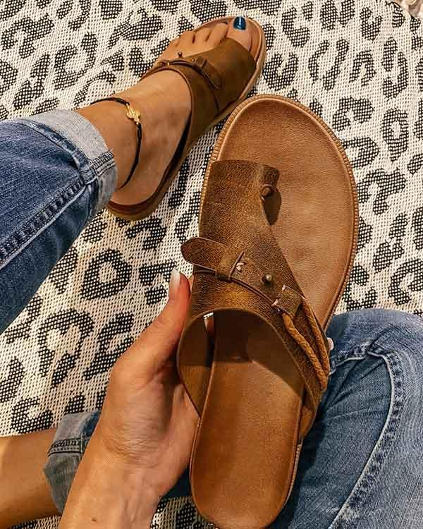 Brown leather sandals with a toe loop and buckle detail, held by a person wearing jeans, on a leopard print rug. Fashionable summer footwear.