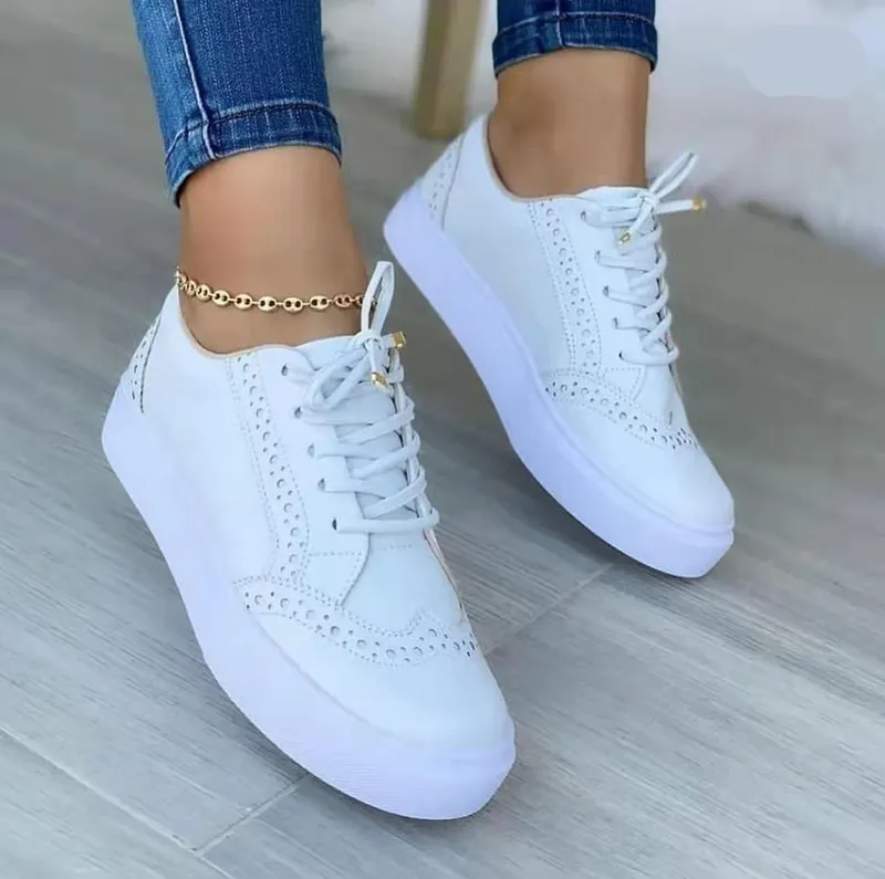 White women's brogue sneakers with perforated design, worn with blue jeans and a gold anklet. Stylish casual footwear for fashion-forward looks.