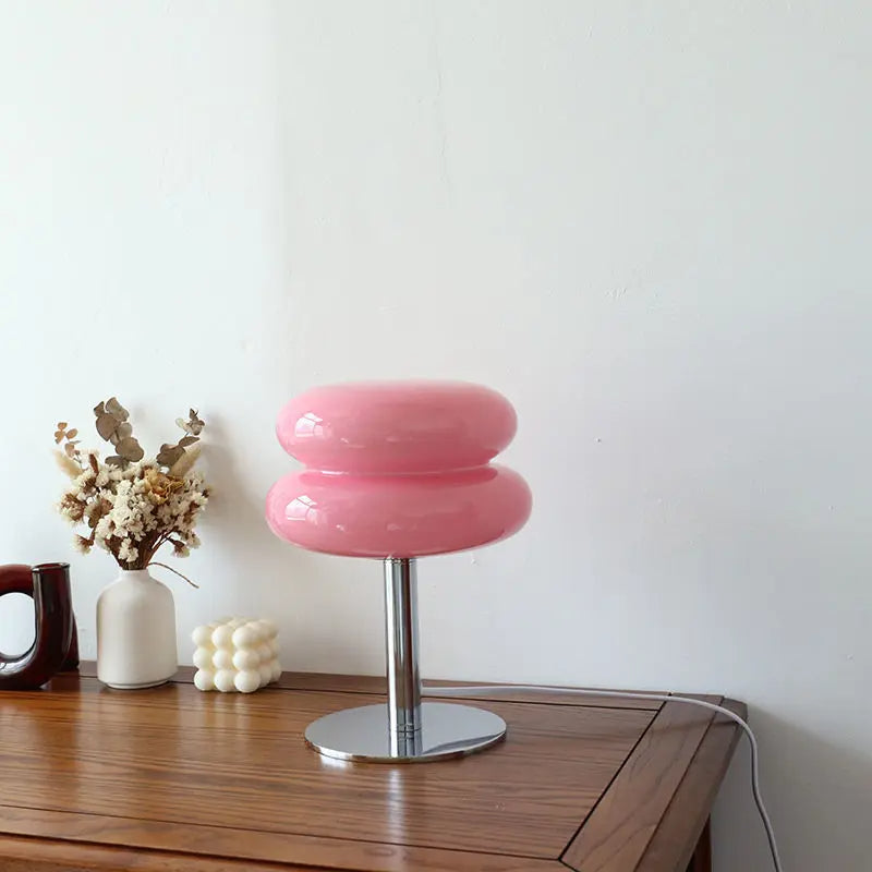 Pink donut-shaped table lamp on a wooden desk, featuring a modern chrome base. Minimalist home decor with dried flowers and a cube candle.