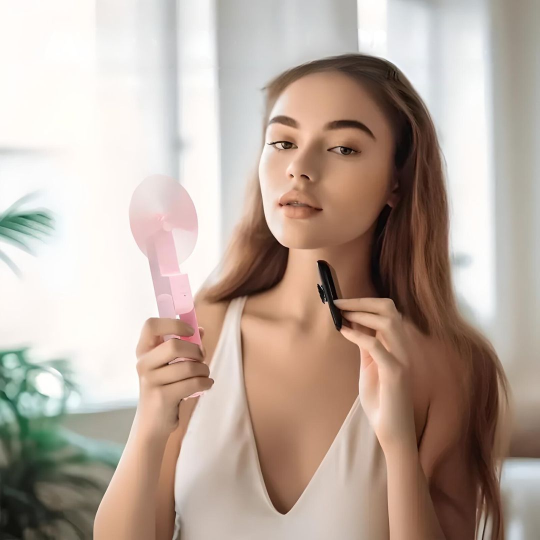 Woman using a pink handheld fan and applying makeup in a bright room; beauty routine, skincare, personal care, lifestyle.