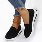 Black slip-on sneakers with white soles, worn with frayed hem jeans and a gold anklet. Stylish women's casual footwear, perfect for everyday wear.