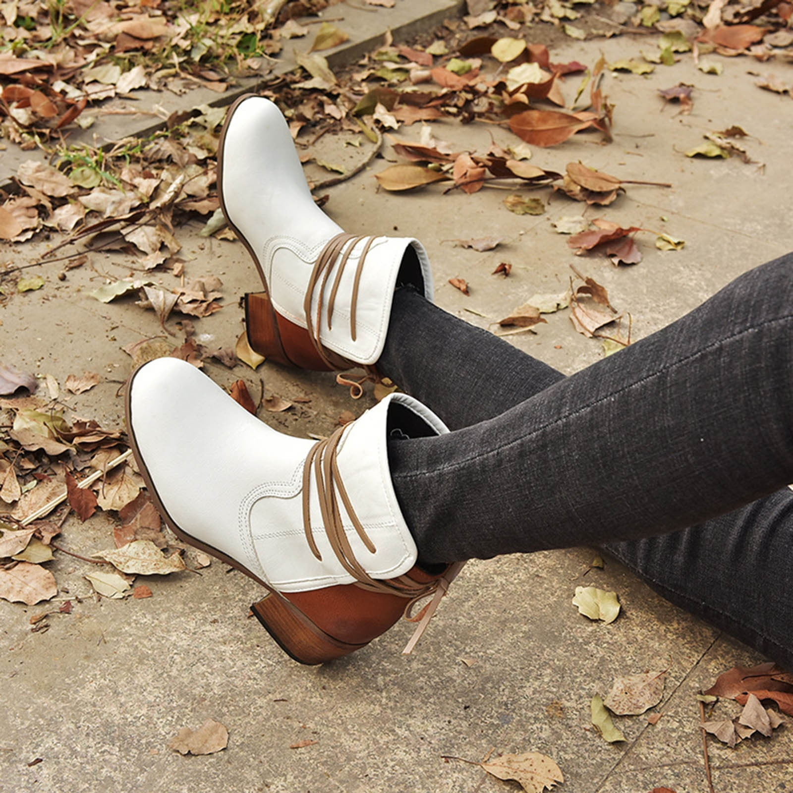 White leather ankle boots with brown laces, worn with dark jeans, surrounded by autumn leaves on a concrete path. Fashionable fall footwear.