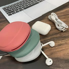 Collapsible silicone lunch box with pink and green lids on wooden desk, next to a laptop, white earbuds, and charging cable. Portable food container.