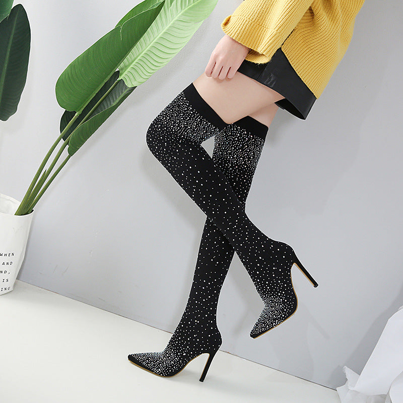 Woman wearing black rhinestone thigh-high stiletto boots, yellow sweater, and black skirt, standing against a gray wall with green plant decor.