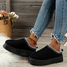 Women's black slip-on platform shoes with white stitching, paired with frayed hem jeans and anklet. Casual footwear fashion on wooden background.