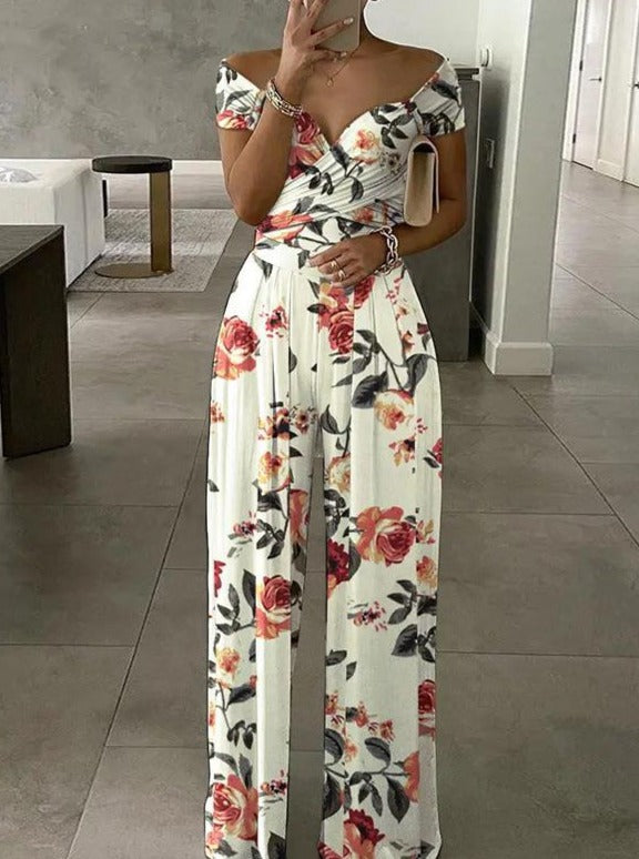 Woman wearing a floral off-shoulder jumpsuit with red and pink roses, holding a clutch, standing in a modern, minimalist interior. Fashionable summer outfit.