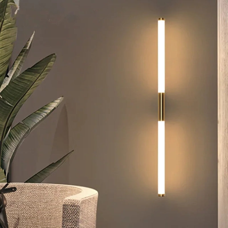 Modern vertical LED wall sconce with warm light, gold accents, and minimalist design, illuminating a contemporary living room with a potted plant.