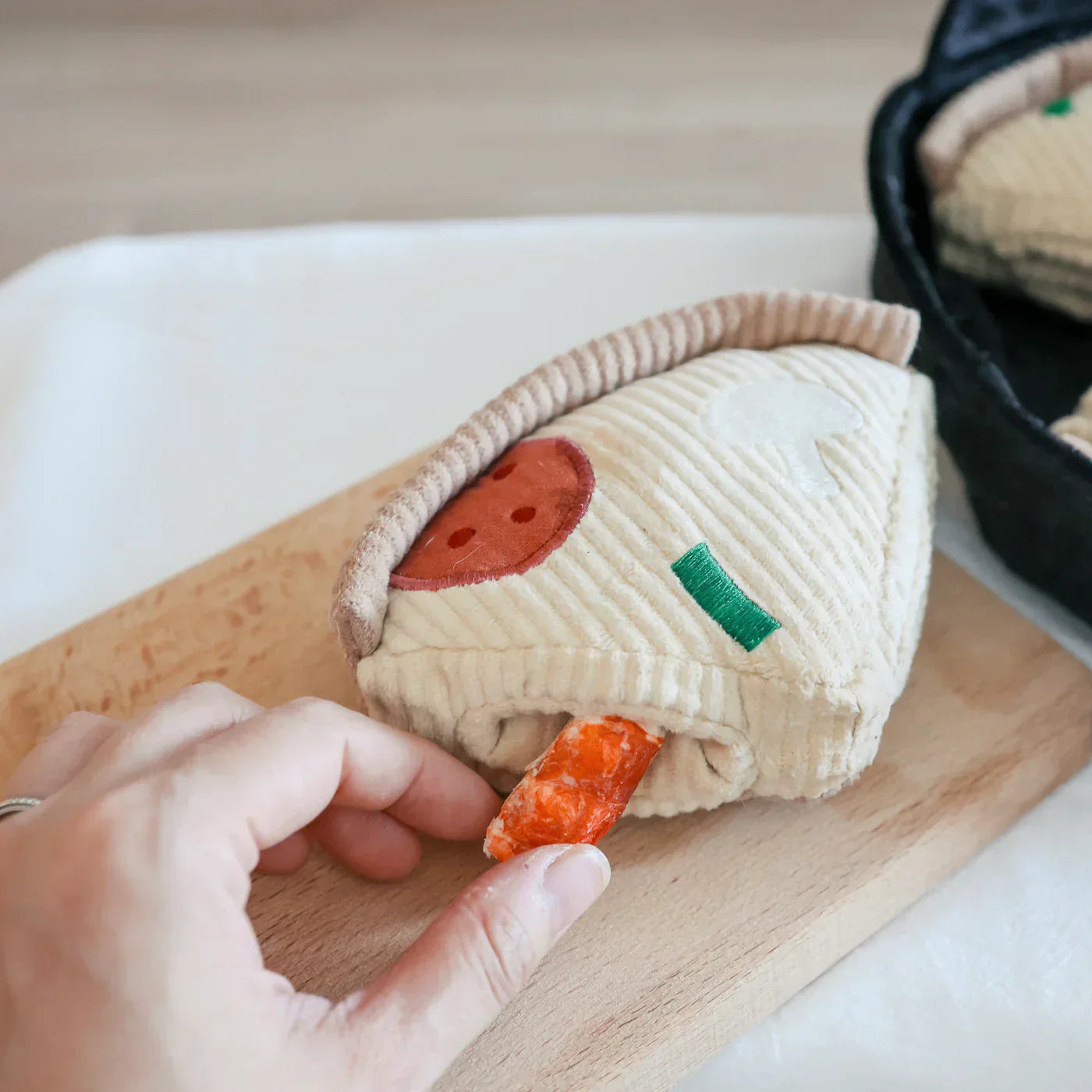 Plush pizza slice toy with embroidered toppings, held by a hand on a wooden board. Soft, textured fabric; ideal for pet play or children's imaginative fun.