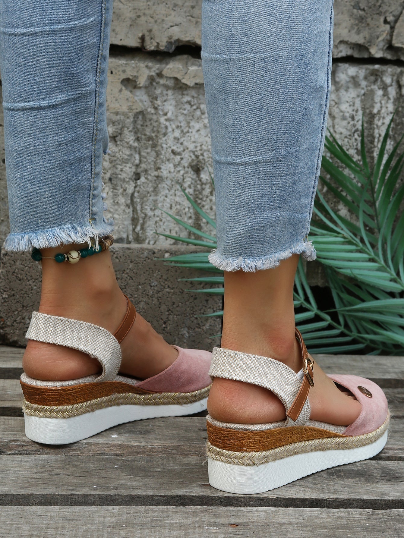 Women's pink wedge sandals with ankle straps and espadrille soles, paired with frayed hem jeans. Fashionable summer footwear on wooden floor.