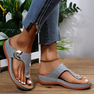 Gray orthopedic sandals with arch support, worn by a person in blue jeans, standing on a wooden floor. Comfortable footwear for women.