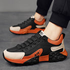 Black and orange athletic sneakers with mesh upper, cushioned sole, and "AIR" branding, worn by a person on a gray surface. Fashionable sports footwear.