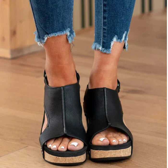 Black open-toe wedge sandals with cork soles, worn with frayed hem jeans on wooden floor. Fashionable women's footwear, summer style.