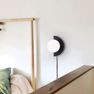 Modern wall sconce with half-moon design, black and white finish, illuminating a minimalist bedroom with wooden bed frame and green leaf pillow.