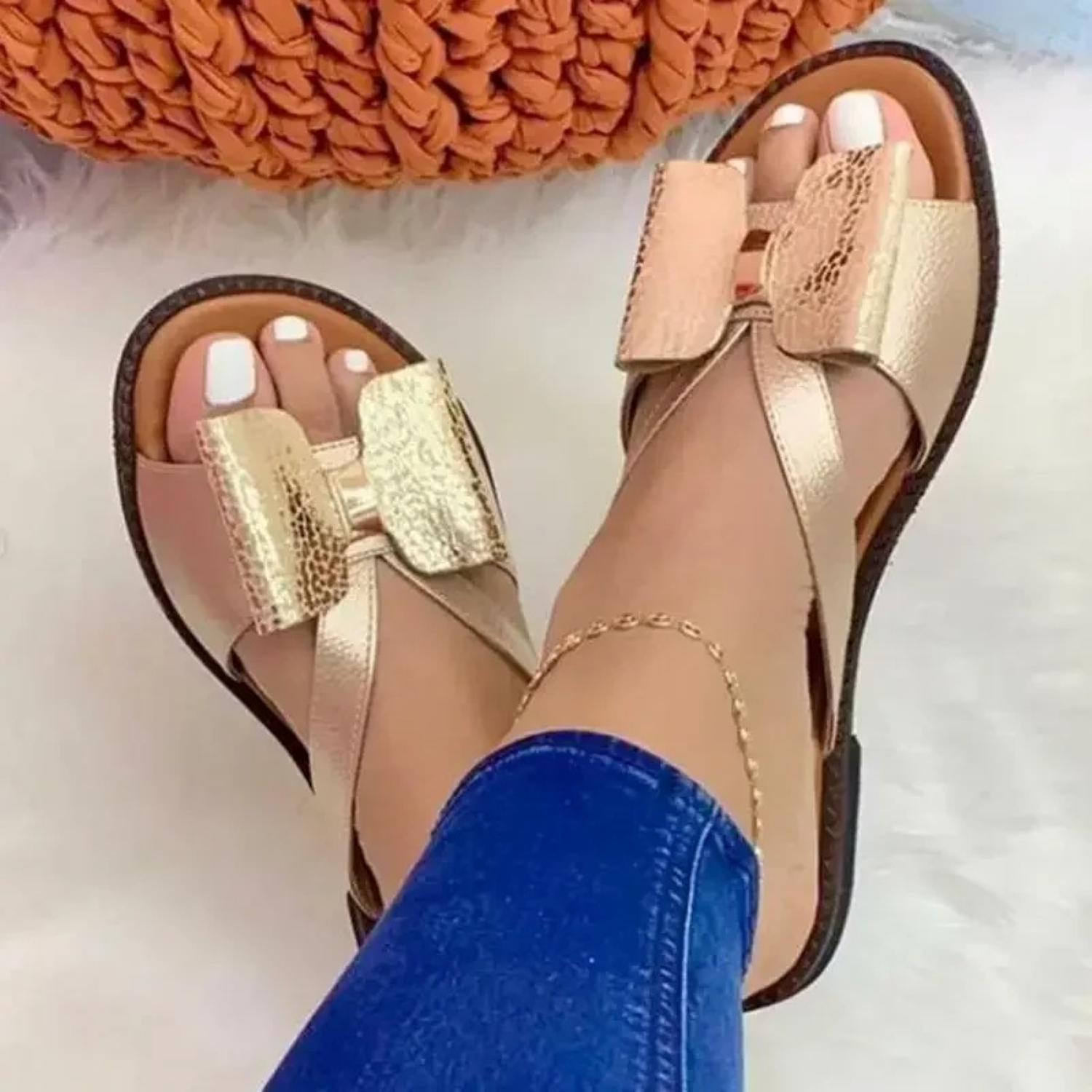 Women's gold bow sandals with crisscross straps, worn with blue jeans. Stylish summer footwear on a white fluffy rug. Fashionable open-toe design.