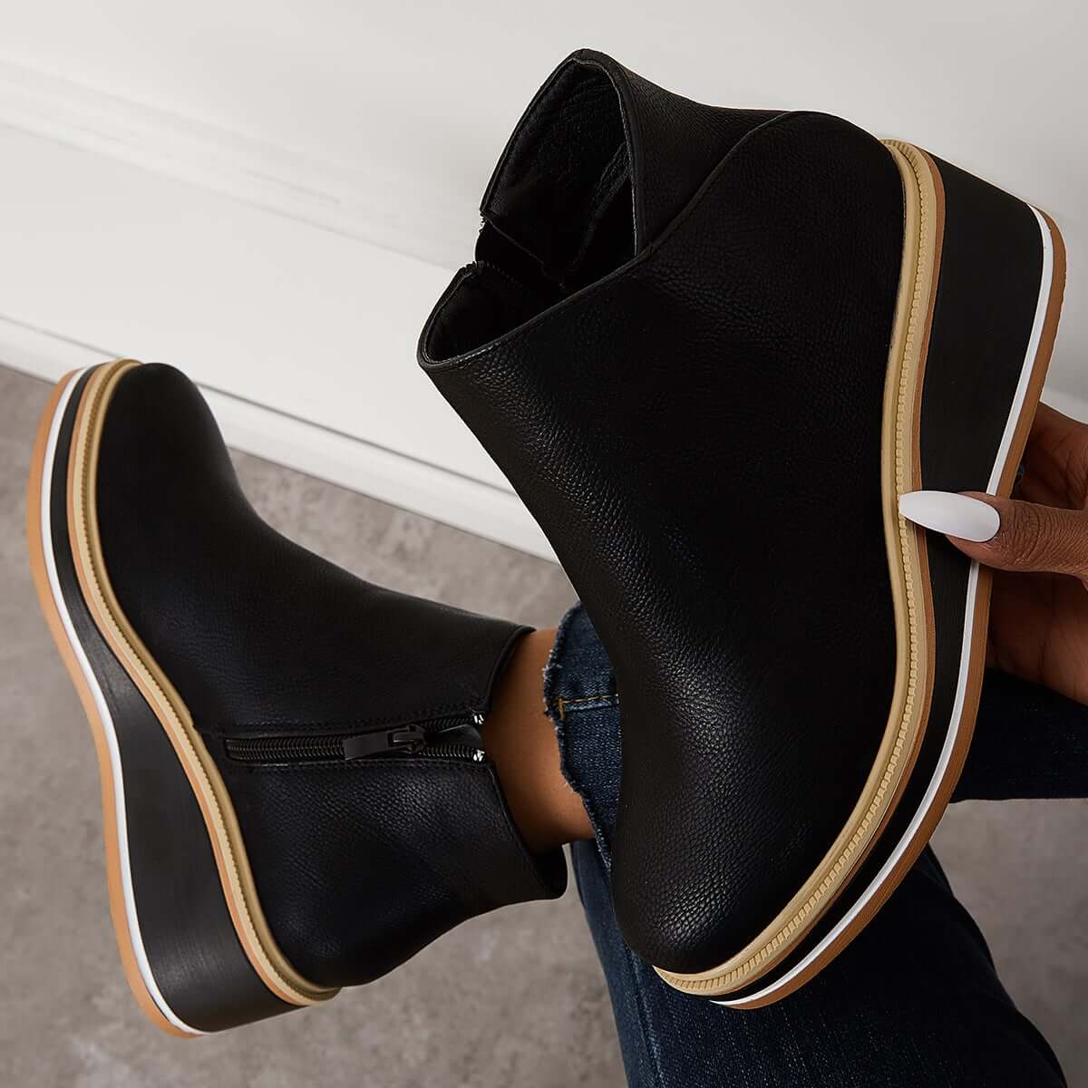 Black wedge ankle boots with side zipper, white sole trim, and tan accents, held by a hand with manicured nails. Fashionable women's footwear.