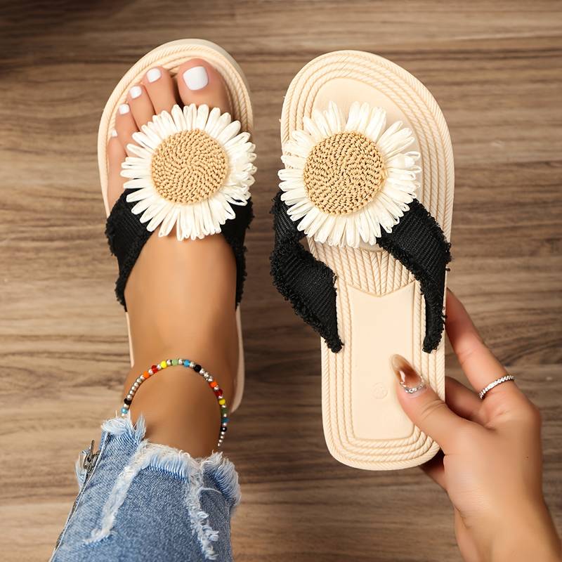 Beige sandals with daisy flower design, black straps, and textured sole on wooden floor. Perfect for summer fashion and casual wear.