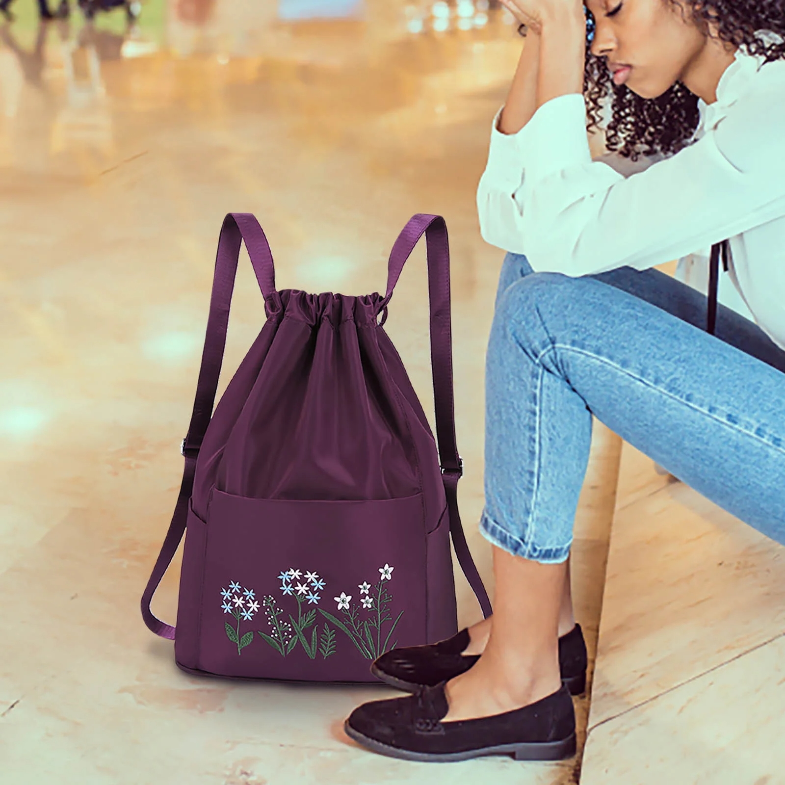 Purple drawstring backpack with floral embroidery, featuring daisies and leaves, on a marble floor next to a seated person. Fashionable and stylish accessory.