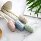 Pastel-colored silicone dish scrubbers with ergonomic handles, displayed on a marble surface near green leaves and white tiles, ideal for kitchen cleaning.