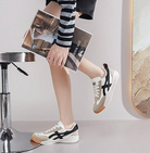 Casual sneakers with black stripes and beige accents, worn by a person in a striped outfit, holding a magazine. Stylish footwear, modern fashion.