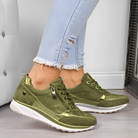 Olive green women's sneakers with white soles, featuring a side zipper and lace-up design, paired with light blue ripped jeans on a wooden floor.