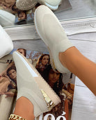 Beige slip-on sneakers with gold heel detail on a wooden floor, reflecting in a mirror. Fashion magazine and gold chain bracelet visible.