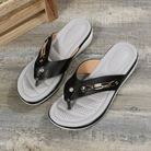 Women's black leather thong sandals with cushioned gray insoles, decorative metal accents, and non-slip soles on a rustic wooden floor background.