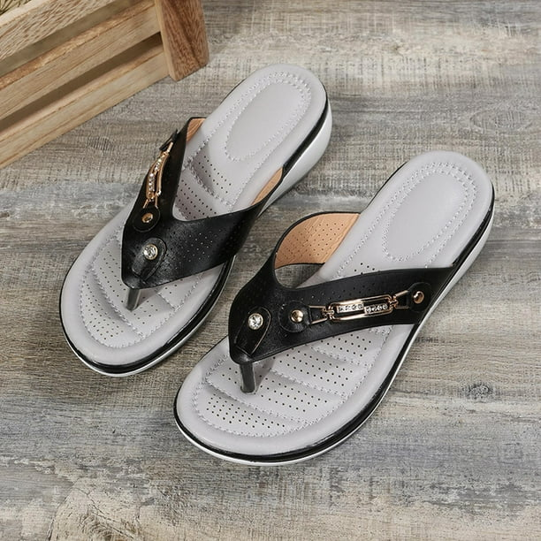 Women's black leather thong sandals with cushioned gray insoles, decorative metal accents, and non-slip soles on a rustic wooden floor background.