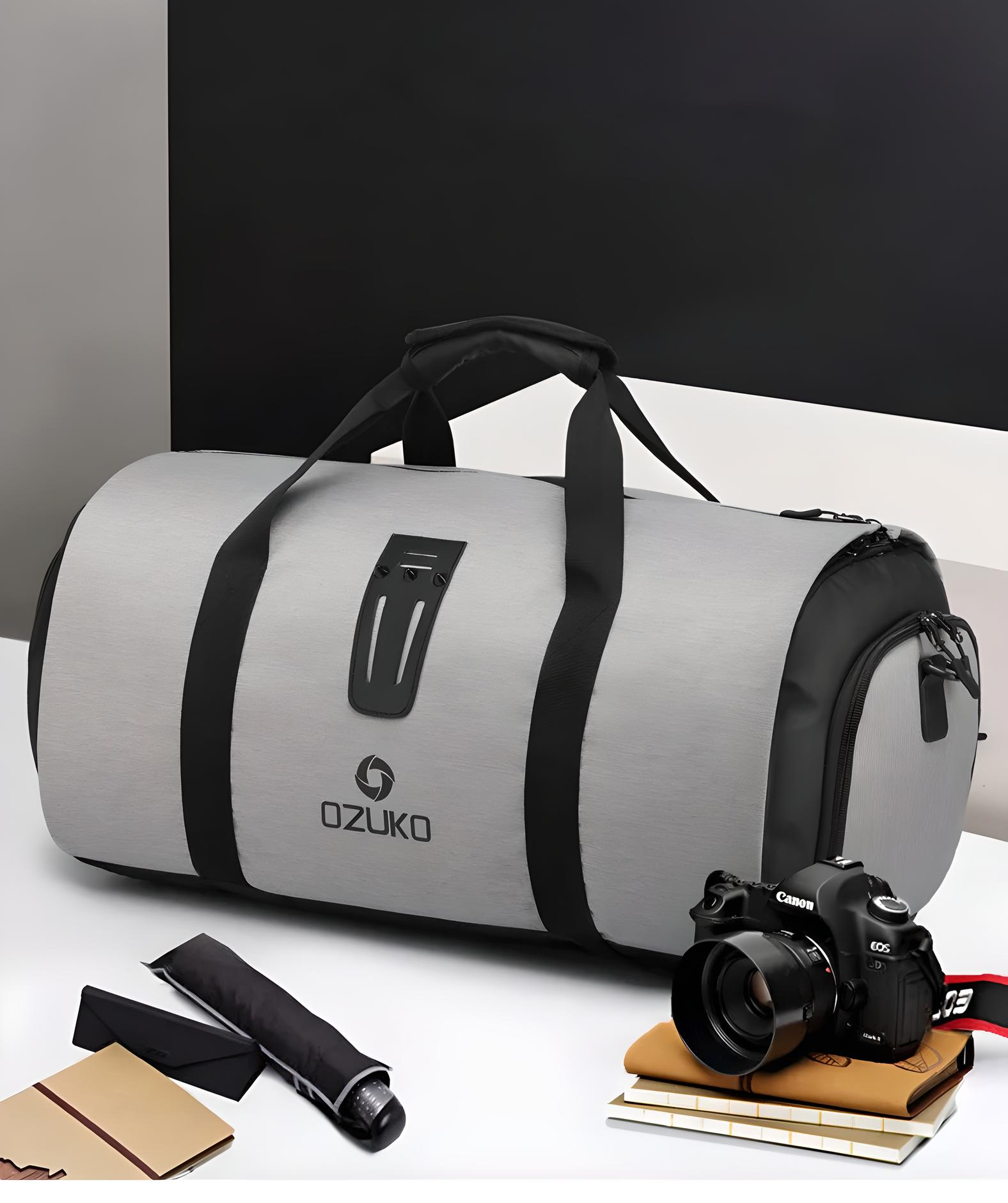 Gray Ozuko duffel bag with black straps on a desk, accompanied by a Canon camera, notebook, and umbrella. Travel gear, stylish luggage.