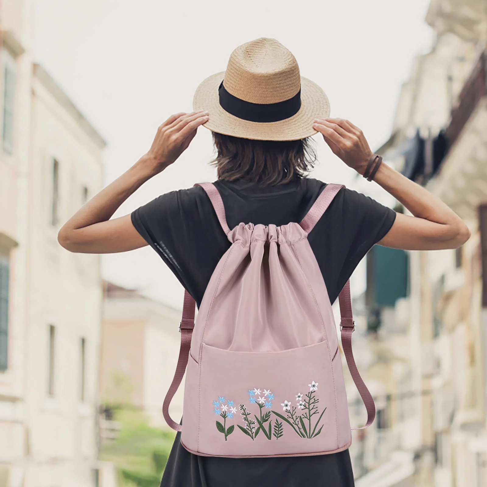 Woman wearing a straw hat and black dress with a pink floral drawstring backpack, standing on a city street. Fashionable urban travel accessory.