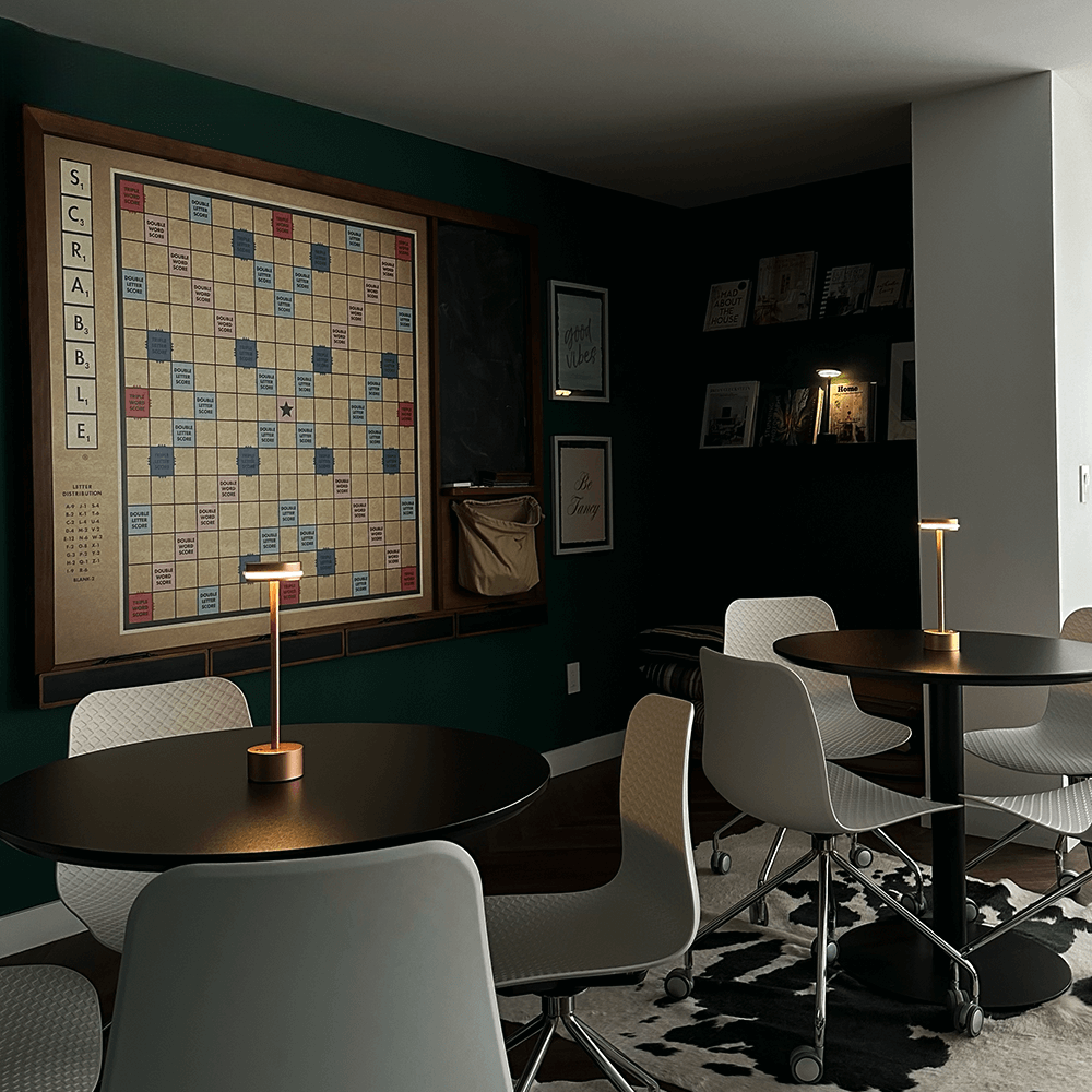 Modern game room with large wall-mounted Scrabble board, round tables, stylish chairs, and ambient lighting. Ideal for board game enthusiasts.