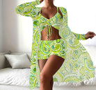 Woman wearing a vibrant green paisley print three-piece outfit, featuring a crop top, shorts, and a long sheer cover-up, standing in a modern living room.