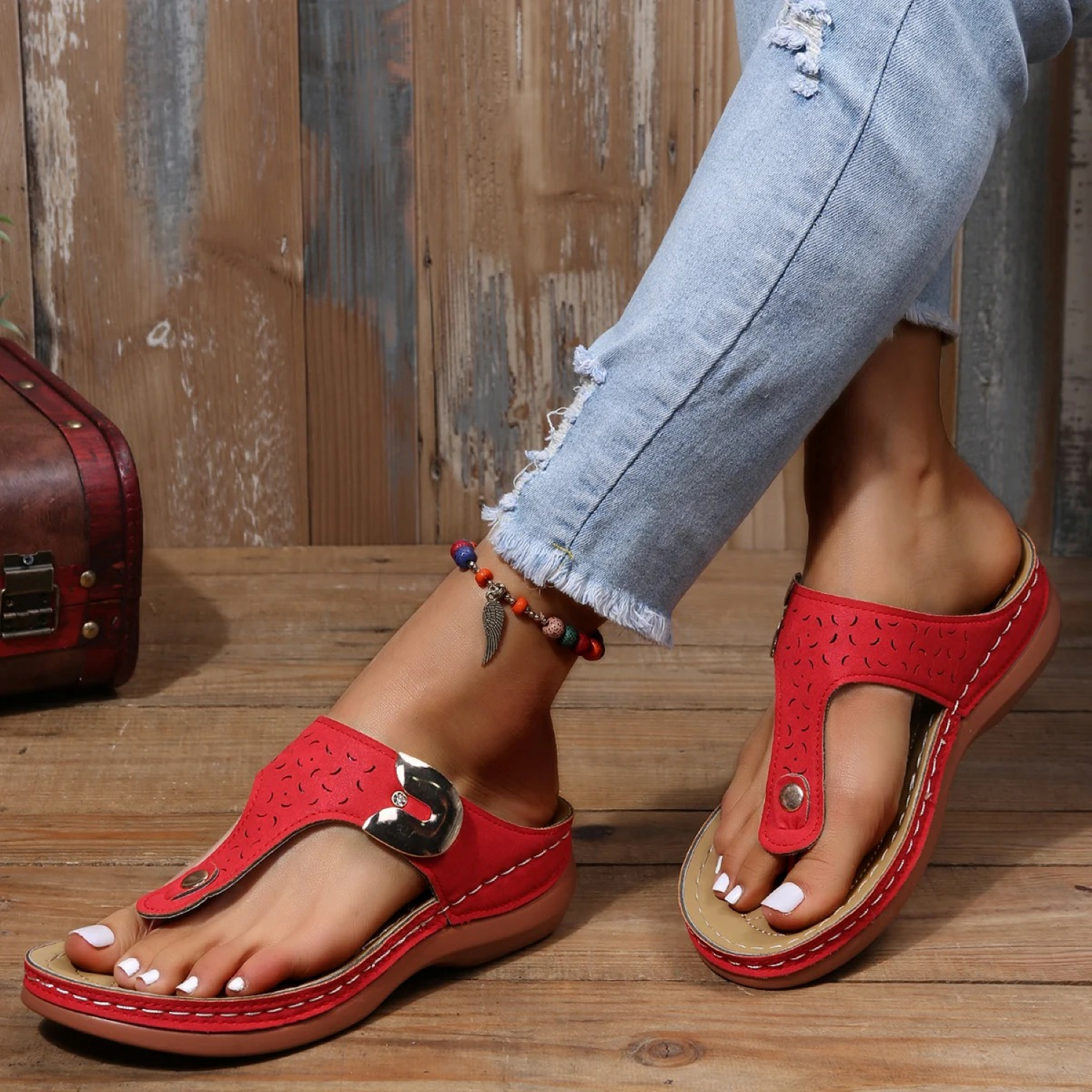 Red leather sandals with buckle detail on woman's feet, paired with ripped jeans. Stylish summer footwear, casual fashion, wooden floor background.