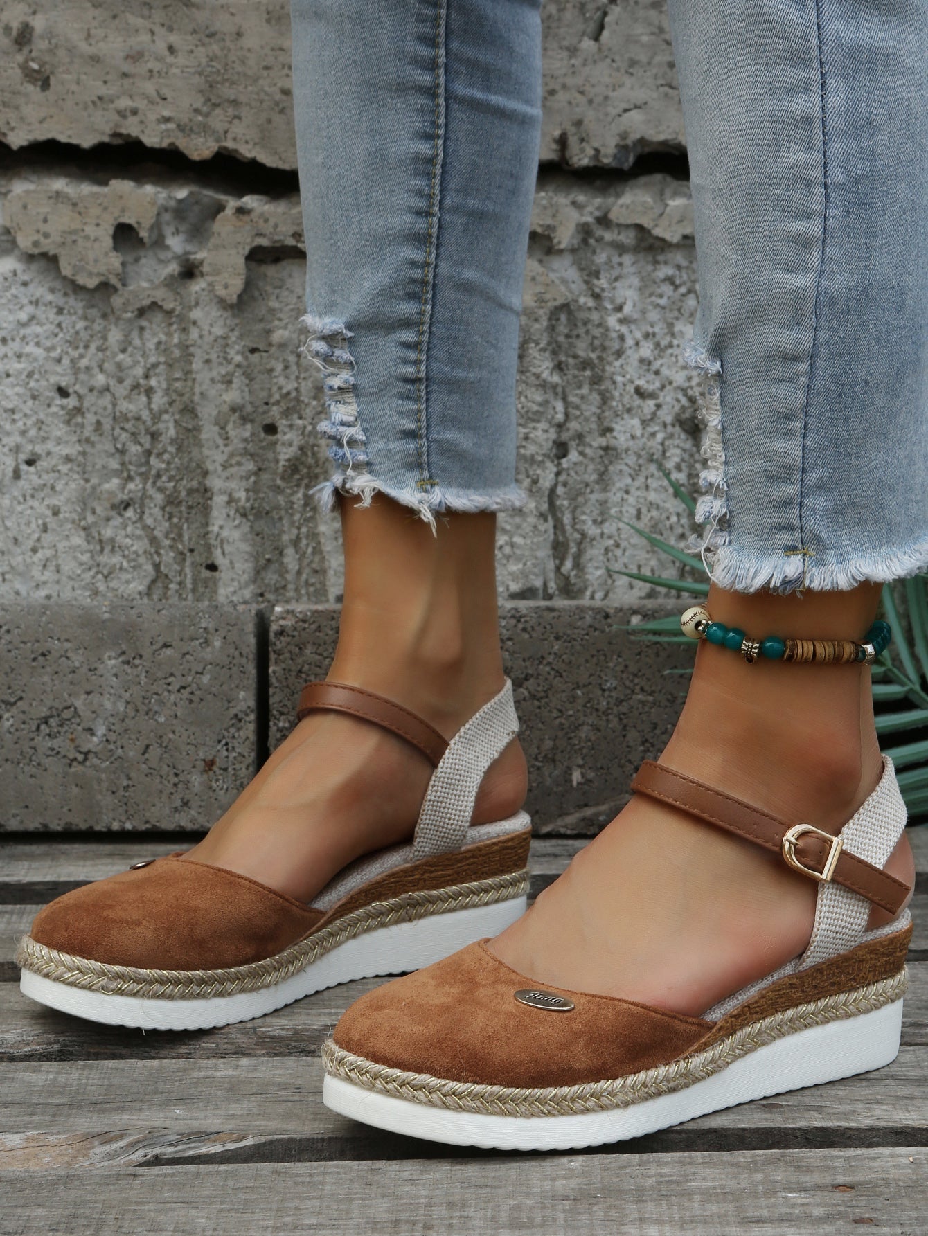 Women's brown suede espadrille wedges with ankle straps, paired with distressed denim jeans. Fashionable summer footwear, casual style.