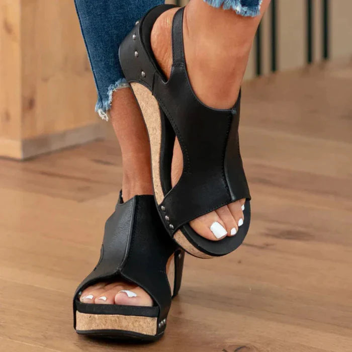 Black open-toe wedge sandals with cork soles, featuring a stylish cutout design and silver rivet accents, worn with frayed hem jeans.