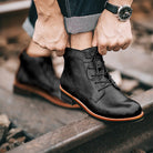 Man tying black leather boots on railroad track, showcasing stylish men's footwear. High-quality, durable boots with brown soles, perfect for casual wear.