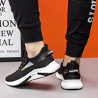 Black and white athletic sneakers with "FASHION" text, worn by a person in black joggers, against an orange and white background. Trendy sports footwear.