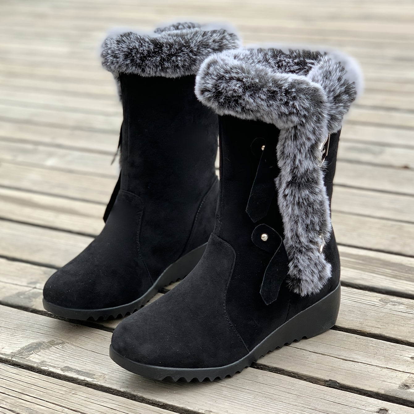 Black suede winter boots with faux fur trim, wedge heel, and decorative tassels on wooden deck. Stylish women's footwear for cold weather.