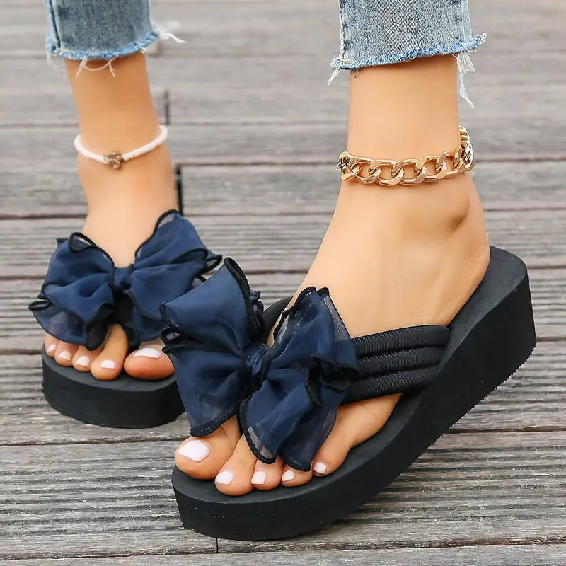 Women's black platform sandals with large navy bows, worn with anklets on wooden deck. Fashionable summer footwear, trendy wedge design.