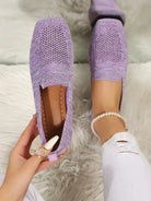 Purple knit slip-on shoes on a woman's feet, featuring a breathable design and paired with white pants and a pearl anklet, on a plush white rug.