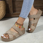 Beige woven platform sandals with crisscross straps on a woman's foot, paired with a colorful anklet. Fashionable summer footwear on wooden floor.