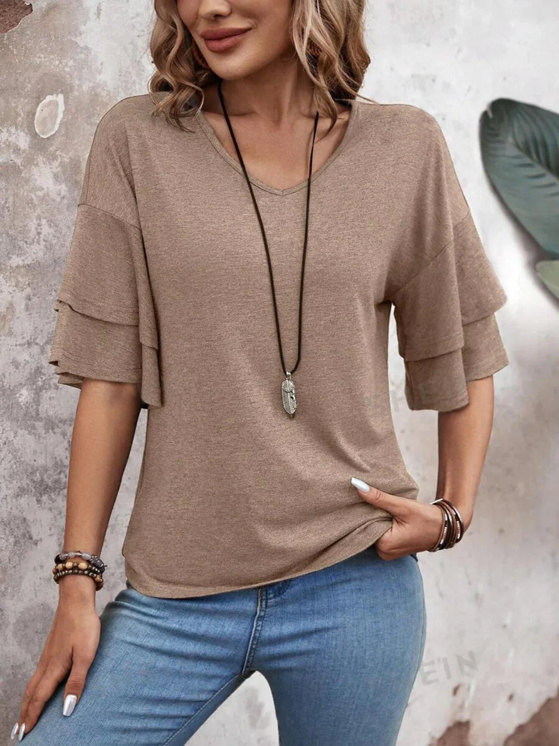 Woman wearing a casual beige short-sleeve top with layered ruffle sleeves, paired with blue jeans and a long necklace, standing against a textured wall.