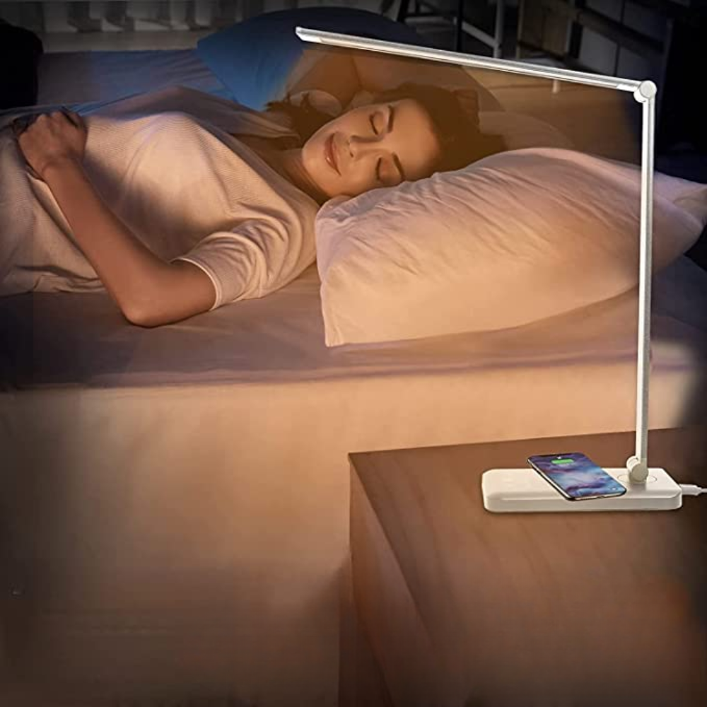 Modern LED desk lamp with wireless charging pad on a bedside table, illuminating a sleeping woman. Ideal for bedroom lighting and smartphone charging.