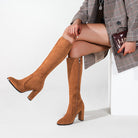 Woman wearing stylish brown suede knee-high boots with block heels, paired with a plaid coat, holding a clutch. Fashionable fall footwear.