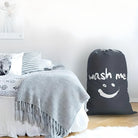 Large black laundry bag with "wash me" text and smiley face, placed beside a cozy bed with gray blanket and decorative pillows in a bright bedroom.